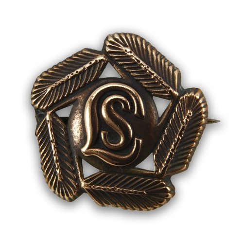 Lottamerkit kuvailtuna: lottien pronssinen hiihto- ja kävelymerkki, jossa havukoristelu reunoilla ja keskellä kirjaimet LS