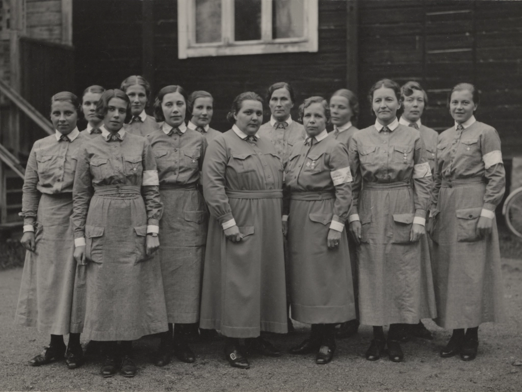 Ryhmää keskellä vinottain seisovilla kahdella lotalla on villakankaiset lottapuvut, muilla puuvillapuvut.