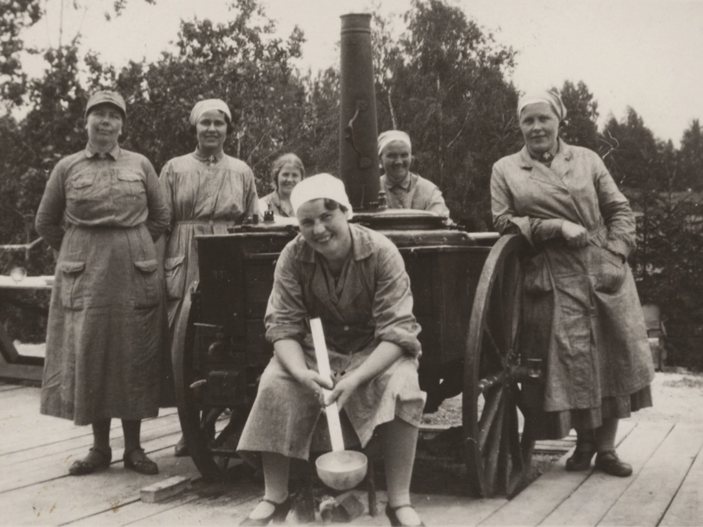 Lottaryhmä harmaissa työasuissa ja päähineissä. Työasu saattoi olla esiliina- tai työtakkimallinen.